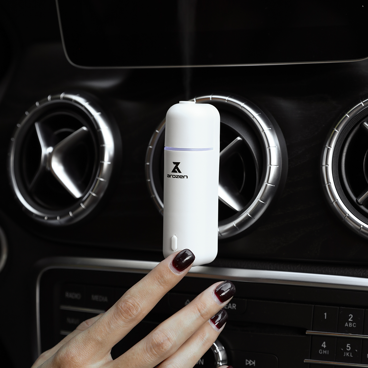 
                  
                    A hand with dark nail polish touches a white AURA X1 CAR SCENT DIFFUSER clipped to a car air vent, adding elegance and fragrance oils to the interior. The dashboard and control buttons are visible in the background.
                  
                