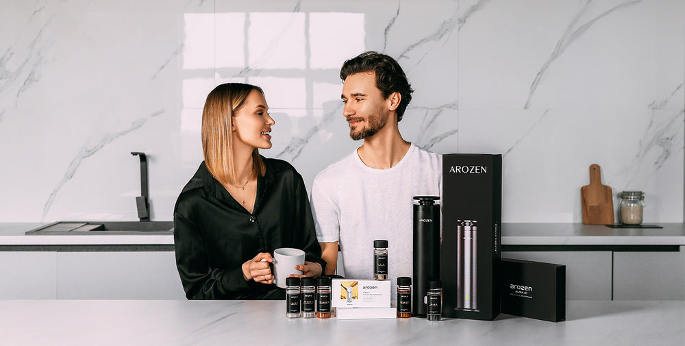 A woman and a man stand in a modern kitchen, smiling at each other. Various Arozin-branded electric scent diffuser products are displayed on the countertop in front of them. The woman holds a white mug.