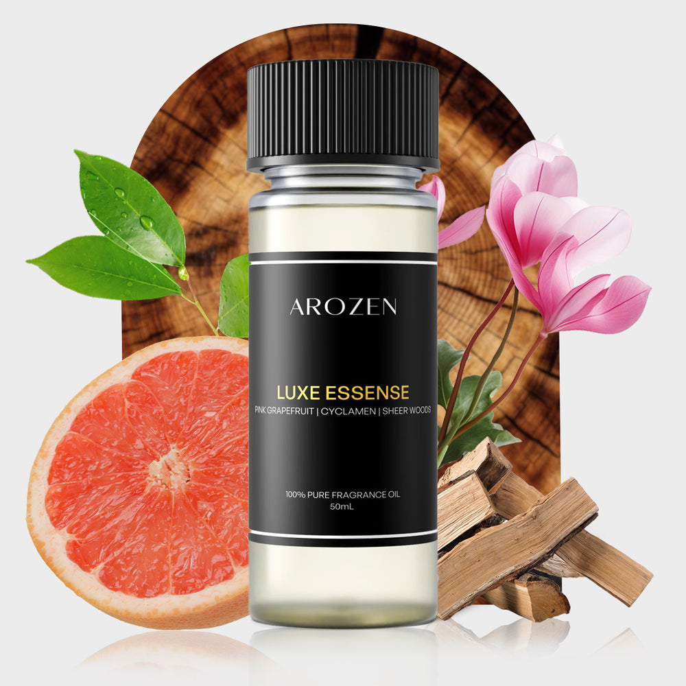 A bottle of Arozen LUXE ESSENCE is shown with grapefruit, green leaves, cyclamen flowers, wood pieces, and a tree slice behind it. The label features pink grapefruit, cyclamen, and sheer woods notes.