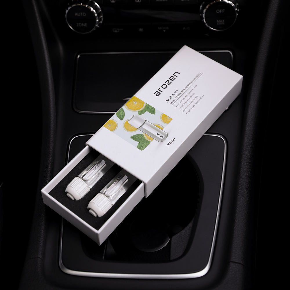 A partially open Arozen box with two small bottles labeled OCEAN - AURA X1 FRAGRANCE OIL REFILL rests on a cars center console. Lemon artwork suggests a fresh, citrus-scented product designed for elegant, coastal-inspired ambience.