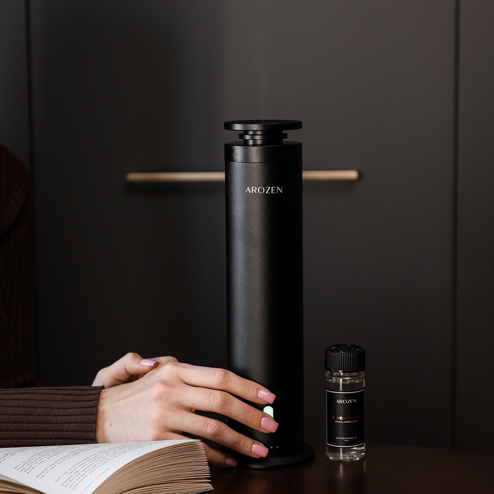 
                  
                    A hand with pastel-painted nails rests on the Arozen EON PRO ELECTRIC SCENT DIFFUSER, a black waterless device set on a dark table next to fragrance oils and an open book, all against a dark background.
                  
                