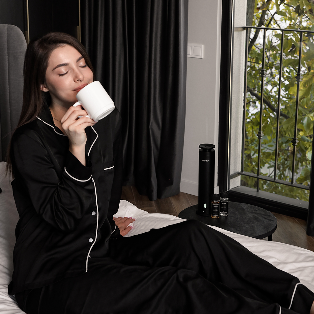 
                  
                    A woman in black pajamas sits on a bed, eyes closed, sipping from a white mug. Beside her, the Arozen EON PRO SCENT DIFFUSER and bottles of fragrance oils rest on a small table by the window framing lush green leaves.
                  
                