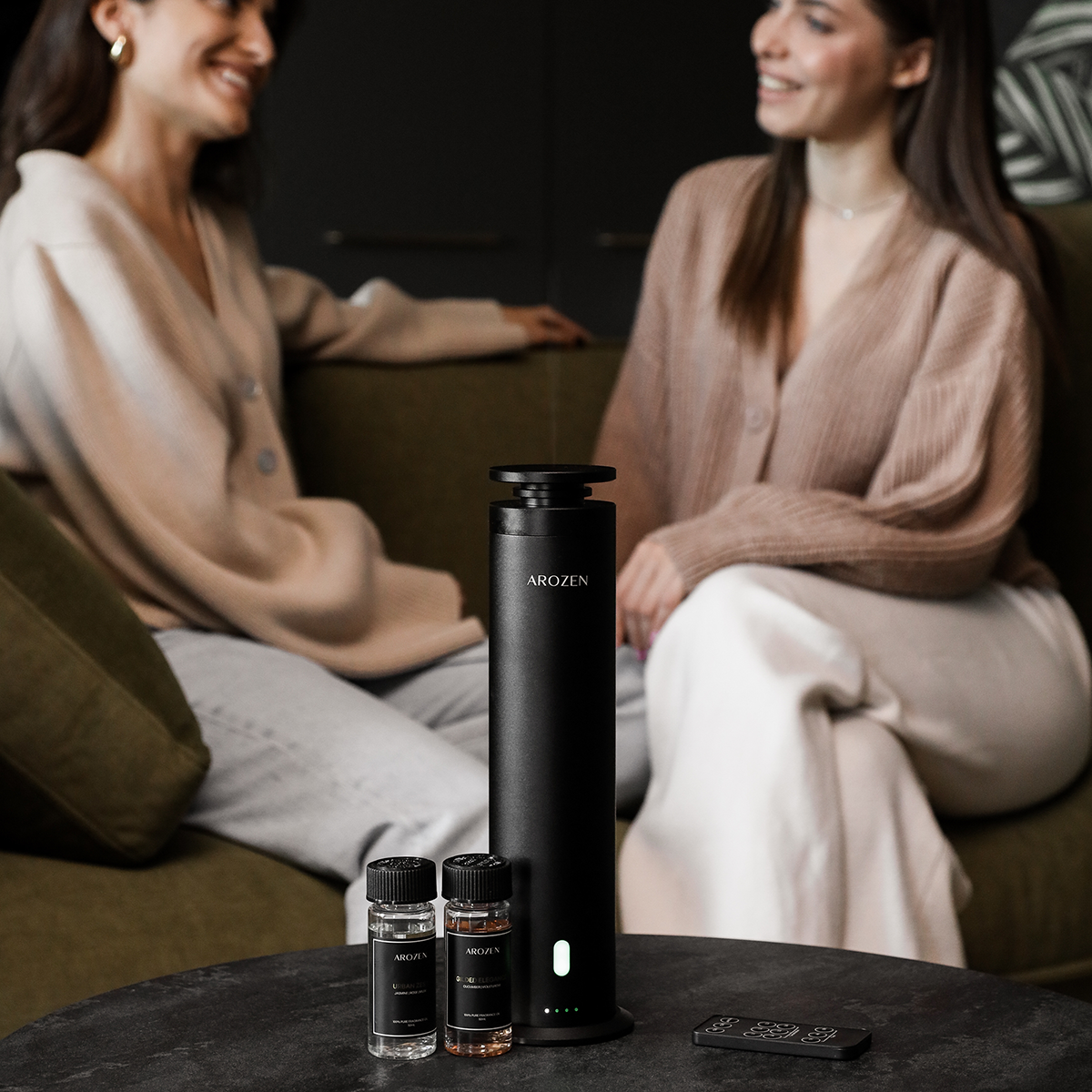 
                  
                    Two women chat on a sofa in the background. On a round table in front, an Arozen EON PRO ELECTRIC SCENT DIFFUSER, two fragrance oils, and a remote control are displayed.
                  
                