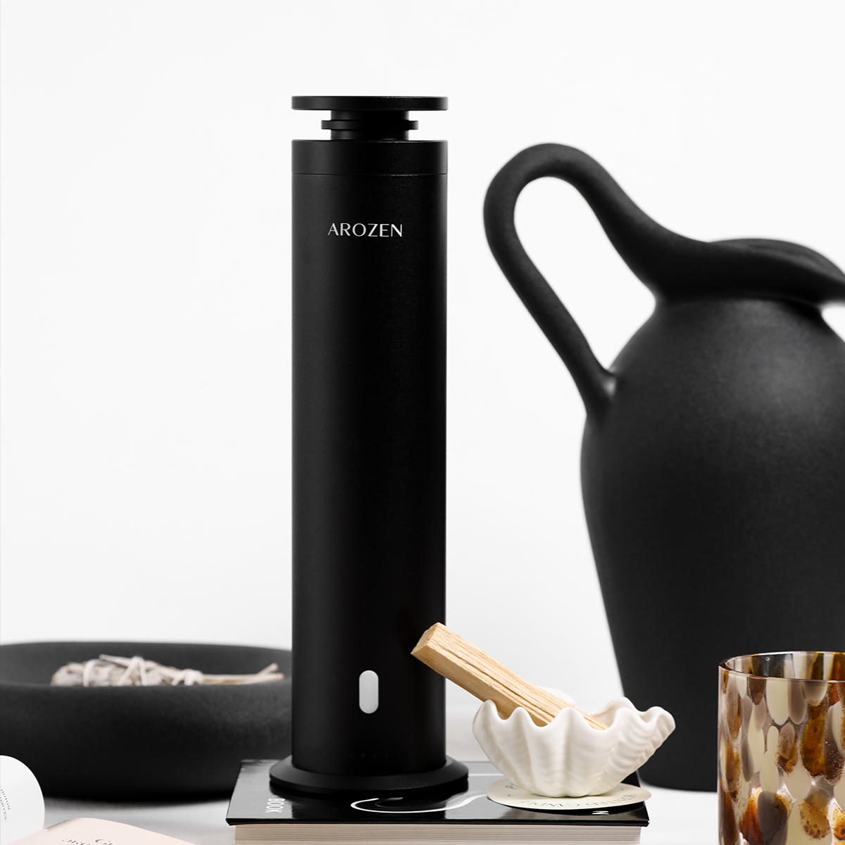 
                  
                    A sleek black cylindrical EON PRO ELECTRIC SCENT DIFFUSER by Arozen stands on a table near a black ceramic pitcher, incense sticks, candle, glass, and an open book—all arranged in minimalist style.
                  
                